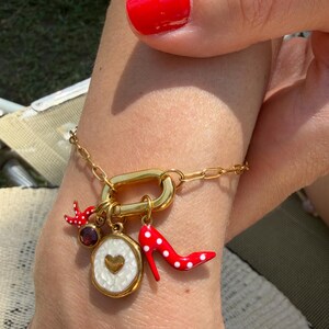 May include: A gold-toned chain bracelet featuring a large oval link adorned with three charms: a small red bird, a round white enamel pendant with a central gold heart, and a red high-heeled shoe charm with white polka dots and a dark red stone.
