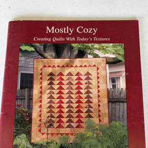 May include: A book titled "Mostly Cozy" with the subtitle "Creating Quilts With Today's Textures." The cover features a quilt with red and brown triangle patterns. The book is framed in a dark red border, with the words "Calico Printworks" at the bottom.