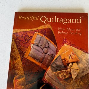 May include: A book titled "Beautiful Quiltagami" with the subtitle "New Ideas for Fabric Folding" is displayed. The cover features images of folded fabric art in shades of orange, brown, and purple. The author's name, Mary Jo Hiney, is also visible.