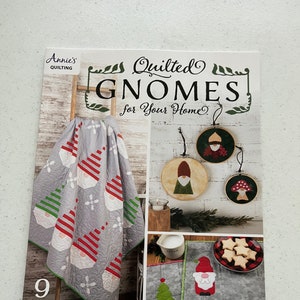 May include: A book cover titled "Quilted Gnomes for Your Home" by Annie's Quilting. The cover features a gray quilt with gnome designs, along with other gnome-themed decorations. The book promises 9 whimsical designs.