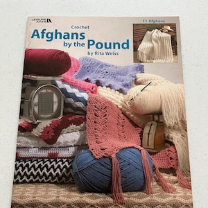 May include: A book cover titled "Afghans by the Pound" by Rita Weiss, featuring various crochet afghans and yarn. The cover includes a stack of folded blankets, balls of yarn, and a scale. The book is published by Leisure Arts.
