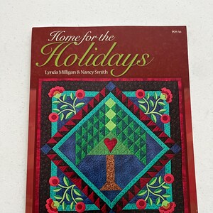 May include: A book cover titled "Home for the Holidays" with a quilt design. The quilt features a tree with a heart, surrounded by floral patterns in red, green, and blue. The cover is framed in a dark red border.