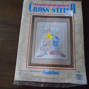 May include: A counted cross-stitch kit featuring a framed image of two children in a room, with the text "WHO STOLE THE COOKIES?" The kit includes a color graph, a 24 Kt. gold plated needle, and instructions. The frame size is 11 x 14 inches.