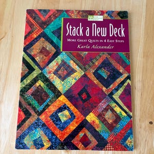 May include: A softcover instructional book titled Stack a New Deck: More Great Quilts in 4 Easy Steps by Karla Alexander. The cover features a vibrant, multi-colored quilt pattern with geometric diamond shapes in shades of red, blue, and green.