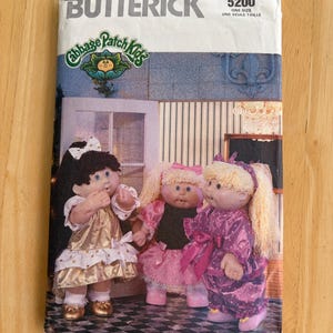 May include: A vintage Butterick sewing pattern featuring Cabbage Patch Kids dolls. The pattern cover displays three dolls in various outfits, with the Butterick logo and pattern number 5200 at the top. The dolls are dressed in gold, pink, and purple outfits.
