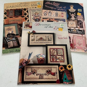 May include: A collection of cross-stitch pattern books featuring gardening themes. The books showcase framed cross-stitch designs with floral, angel, and garden tool motifs. Titles include "Gardening Angel" and "Blessings of the Earth."