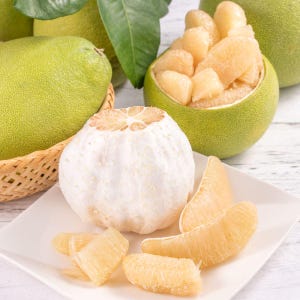 May include: A still-life image of pomelos, showcasing the fruit's various stages. Whole green pomelos are in the background, with one in a woven basket. A peeled pomelo sits on a white plate with several segments. The peeled fruit is white, and the segments are a pale yellow.