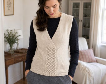 May include: A cream-colored sleeveless knit vest featuring a V-neckline and a central vertical cable-knit panel. The garment is worn over a dark navy long-sleeved shirt and paired with a dark gray skirt, showcasing a textured, layered appearance.