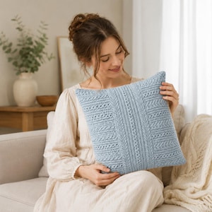 May include: A square light blue throw cushion featuring a textured knit pattern with vertical columns of intricate lace and cable stitches. The cushion has a uniform soft blue colour and a detailed, repeating geometric design across the entire surface.