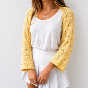May include: A pale yellow knit bolero shrug featuring an openwork diamond pattern and three-quarter length sleeves, worn over a plain white scoop-neck top and a white pleated skirt. A silver-toned heart pendant necklace is worn around the neck.