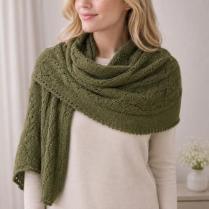 Peut inclure: Un châle tricoté vert olive présentant un motif en dentelle complexe avec un bord festonné délicat. Le châle est drapé sur un haut uni à manches longues de couleur claire, mettant en valeur la texture détaillée et le design ajouré du tricot.