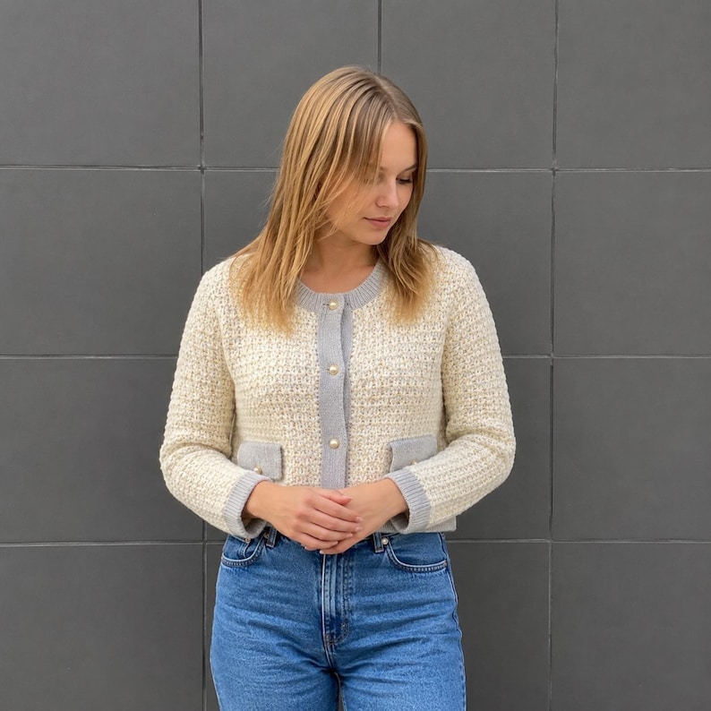 May include: A cream-colored knit cardigan featuring a textured weave, a round neckline, and long sleeves. The garment includes a grey button-down placket with gold-toned buttons and two small front pockets with grey trim, paired with blue denim jeans.