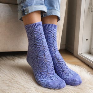 May include: A pair of light purple knit socks featuring an intricate, repeating leaf-like lace pattern throughout the leg and foot, finished with a ribbed cuff at the top and a solid knit texture on the toe and heel sections of the garment.