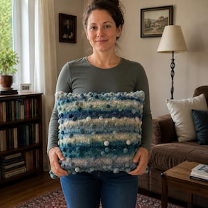 May include: A square knit throw cushion featuring a variegated colour palette of blue, teal, green, and white. The surface is decorated with numerous small, raised pom-poms in matching shades, creating a textured, multi-dimensional appearance.