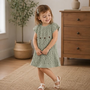 Könnte beinhalten: Ein salbeigrünes Häkelkleid mit einem zarten, durchbrochenen Blattmuster, kurzen Ärmeln und einem Rundhalsausschnitt. Das Kleid ist mit kleinen braunen Zierakzenten im Brustbereich versehen und wird mit weißen Sandalen und einer Haarspange getragen.