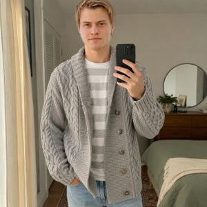 Puede incluir: Un cárdigan de punto trenzado gris claro con cuello de chal y cuatro botones, usado sobre una camiseta de rayas horizontales blancas y grises y jeans azul claro. El tejido presenta un patrón texturizado en las mangas y paneles frontales.