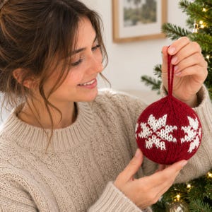 Pode incluir: Um enfeite de Natal tricotado, de formato redondo, em vermelho profundo com um padrão de flocos de neve brancos. A decoração esférica apresenta uma textura de lã e inclui uma alça de lã vermelha a condizer no topo para pendurar.