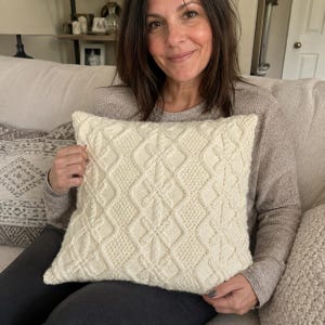 May include: A square, off-white knit throw pillow cover featuring a textured cable knit pattern with diamond shapes and central cross motifs. The fabric has a thick, yarn-based construction with a consistent cream colour throughout the entire surface.