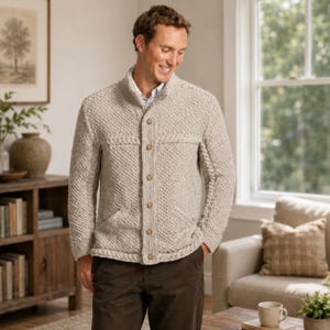 Puede incluir: Un cárdigan de punto beige con un patrón de tejido diagonal texturizado, cuello alzado y cierre frontal con cinco botones de color marrón claro. La prenda tiene mangas largas y dos bolsillos delanteros, sobre una camisa de cuello claro.