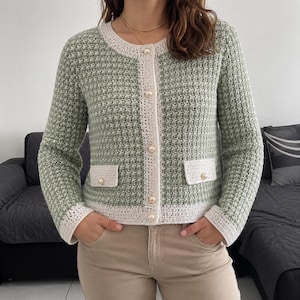 May include: A sage green long-sleeved cardigan with a textured knit pattern, featuring a cream-colored trim along the round neckline, front placket, cuffs, and two front pockets. The garment is fastened with five round, cream-colored pearl buttons.