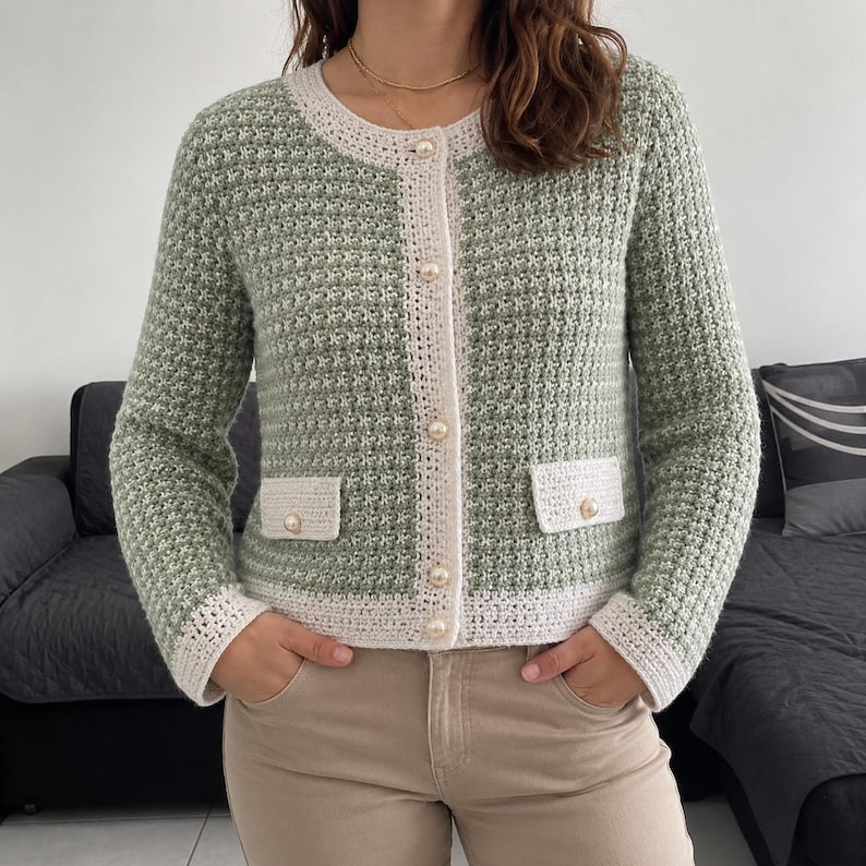 May include: A sage green long-sleeved cardigan with a textured knit pattern, featuring a cream-colored trim along the round neckline, front placket, cuffs, and two front pockets. The garment is fastened with five round, cream-colored pearl buttons.
