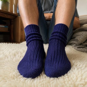 Puede incluir: Un par de calcetines azul marino oscuro con una textura de punto acanalado en todo el pie y la pierna. Los calcetines tienen un diseño holgado y fruncido alrededor de los tobillos, creando una apariencia suave y superpuesta en azul.
