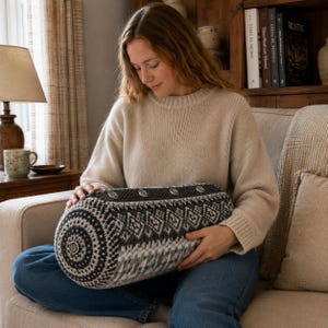 May include: A cylindrical bolster pillow featuring a complex knit pattern in shades of charcoal, gray, and white. The pillow is accented with a row of four metallic buttons along the top edge and a single matching button centered on the circular end panel.