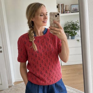 May include: A short-sleeved, coral-colored knit sweater featuring a repeating vertical leaf-like lace pattern. The garment has a round neckline and a ribbed hem, worn over a dark blue collared shirt, showcasing a textured, open-knit construction.
