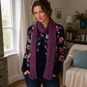 Könnte beinhalten: Ein langer, violetter Strickschal mit gerippter Textur und dekorativen Fransen an beiden Enden. Der Schal ist um den Hals drapiert und hängt über einer dunkelblauen Strickjacke mit Blumenmuster, die über einem blau-schwarz gestreiften Oberteil getragen wird.