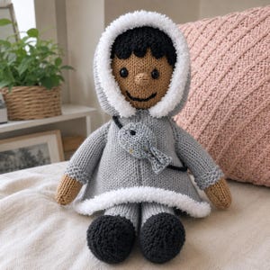 May include: A handcrafted knit doll with dark skin, black hair, and black eyes. The doll wears a light gray hooded coat with white fuzzy trim and a small gray fish-shaped bag worn across the chest. The doll has black shoes and tan hands.
