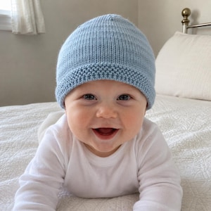 May include: A light blue knit baby hat featuring a textured garter stitch brim and a smooth stockinette stitch crown. The hat is constructed from soft yarn and provides a snug fit, showcasing a classic design suitable for infants and young children.
