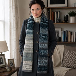 May include: A long knit scarf featuring a gradient of blue, grey, and white colors with intricate geometric patterns. The scarf is worn over a dark charcoal grey wool coat with three visible buttons and a high collar, creating a layered look.