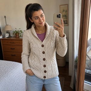 May include: A beige cable-knit cardigan featuring a shawl collar, long sleeves, and six dark brown buttons down the front. The knit pattern includes vertical cable textures and ribbed cuffs, worn over a solid purple shirt and light blue denim jeans.