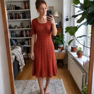 May include: A knee-length, rust-coloured knit dress featuring a vertical leaf-like pattern throughout the fabric, short sleeves, and a rounded neckline. The garment has a fitted bodice that transitions into a slightly flared skirt silhouette.