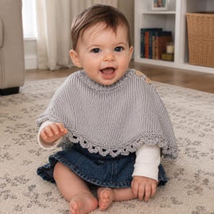 May include: A light gray knitted poncho featuring a ribbed neckline, two small wooden buttons on the shoulder, and a decorative scalloped edge along the bottom hem. The garment is worn over a white long-sleeved shirt and a dark blue denim skirt.