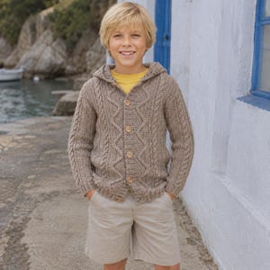 May include: A beige cable-knit hooded cardigan featuring a button-down front with four light brown buttons, long sleeves, and a textured pattern. The cardigan is worn over a plain yellow t-shirt and paired with light beige shorts.
