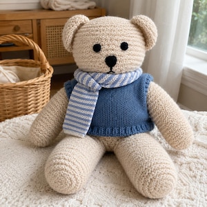 Könnte beinhalten: Ein beigefarbener gehäkelter Teddybär mit einer einfarbigen blauen Strickweste und einem passenden hellblau-weiß gestreiften Schal. Der Bär hat schwarze gestickte Augen und eine schwarze Nase sowie eine strukturierte Garnoberfläche.