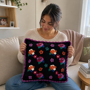 May include: A square black throw pillow featuring a colorful floral pattern with orange, pink, purple, and teal embroidered flowers. The pillow is finished with a decorative purple crocheted scalloped edge trim around the entire perimeter.