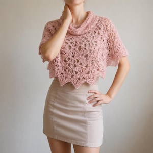 May include: A dusty pink crochet poncho featuring an intricate openwork lace pattern with a scalloped hemline and a draped cowl neckline, paired with a solid light beige mini skirt that has a structured waistband and vertical seam detailing.