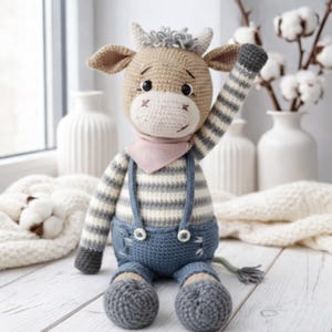 Pode incluir: Boneca de vaca de crochê feita à mão. A vaca tem cabeça, orelhas e chifres castanhos claros. Ela usa uma camisa listrada, macacão azul e um lenço rosa. A vaca está acenando com o braço direito. A boneca está sentada em uma superfície branca.