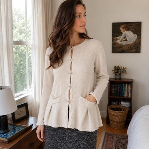 May include: A long-sleeved, cream-colored knit cardigan featuring a round neckline, a button-down front, and two front pockets with pleated detailing at the hem. The garment has a textured knit finish and a slightly flared silhouette at the waist.