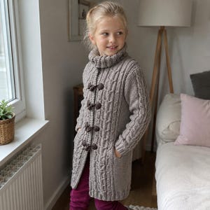 May include: A long-sleeved, beige cable-knit cardigan featuring a high turtleneck collar and four dark brown toggle closures down the front. The garment includes two side pockets and is crafted with a textured, vertical cable pattern.