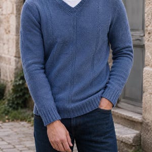 May include: A blue long-sleeve V-neck jumper with vertical ribbed detailing, worn over a plain white crew-neck t-shirt, paired with dark blue denim jeans. The jumper features a textured knit pattern and ribbed cuffs and hem.