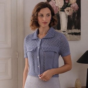 May include: A periwinkle blue short-sleeved knit cardigan featuring a collared neckline, a button-down front with small white floral-shaped buttons, and a combination of openwork lace patterns on the upper chest and vertical panels on the body.