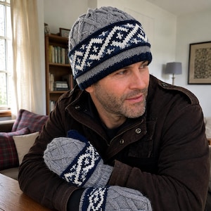 May include: A grey knit beanie and matching mittens featuring a dark blue and white geometric fair isle pattern band. The beanie has a textured cable knit design on the crown, while the mittens display a similar cable knit and patterned cuff.