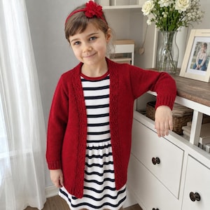 May include: A long-sleeved, open-front red knit cardigan with a textured border along the opening, worn over a white dress featuring horizontal navy blue stripes. A red fabric flower headband is worn on the head, completing the ensemble.