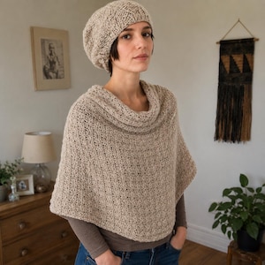 May include: A beige knit poncho with a loose, draped cowl neckline and a matching slouchy knit beanie. The garment features a textured, open-stitch pattern in a neutral tan yarn, providing a lightweight layered look over a long-sleeve shirt.