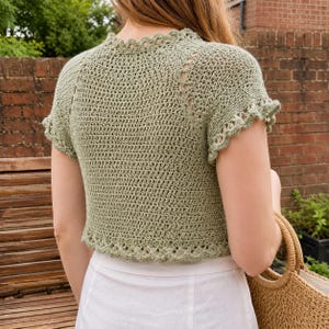 May include: A sage green short-sleeved crochet top featuring a delicate scalloped edge along the neckline, sleeves, and hem. The garment is crafted with a dense, uniform stitch pattern and includes decorative openwork detailing along the raglan seams.