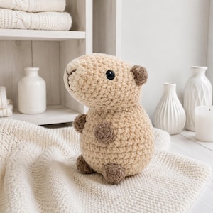 May include: A crocheted capybara plush toy in shades of tan and brown. The handmade stuffed animal has a rounded body, small ears, and a black eye. It sits on a cream-colored knitted blanket, with white vases and a shelf in the background.
