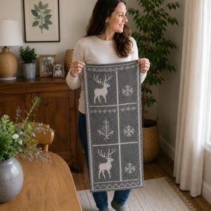 May include: A rectangular grey knit runner featuring a white jacquard pattern of reindeer, snowflakes, and pine trees arranged in a grid. The textile has a decorative border and a consistent knit texture throughout the entire surface of the fabric.